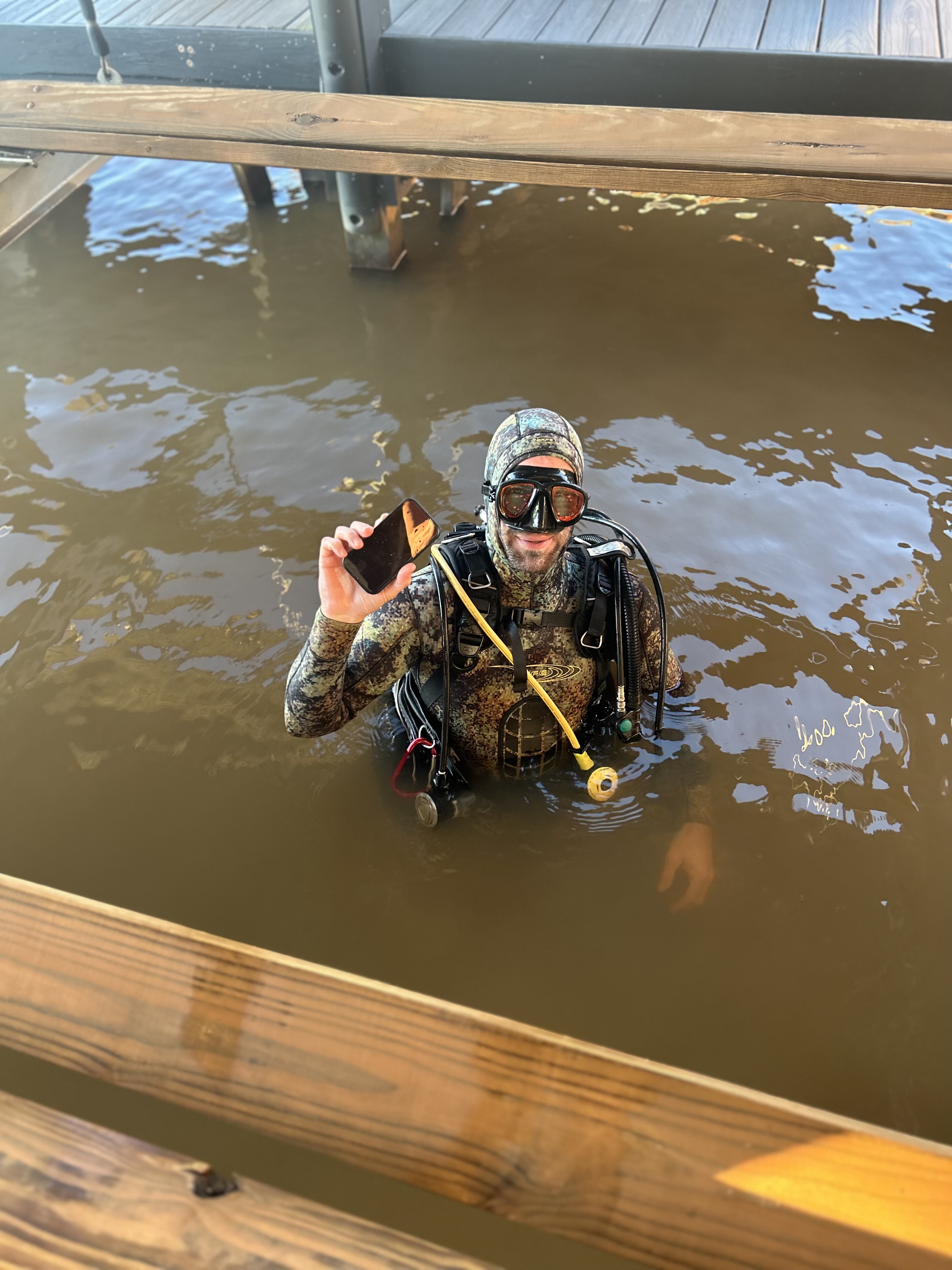 Phil surfacing in a camo wetsuit at a Lake Norman dock, holding a recovered iPhone