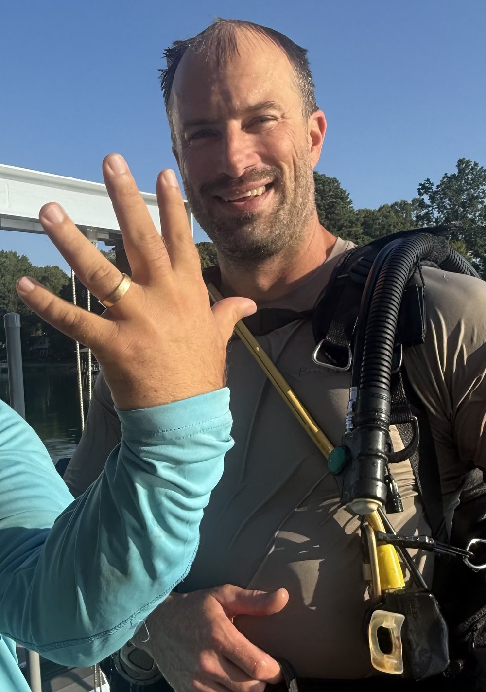Phil in scuba gear, grinning beside a boat lift on Lake Norman, as the owner holds up a hand with the recovered gold wedding band back on the ring finger